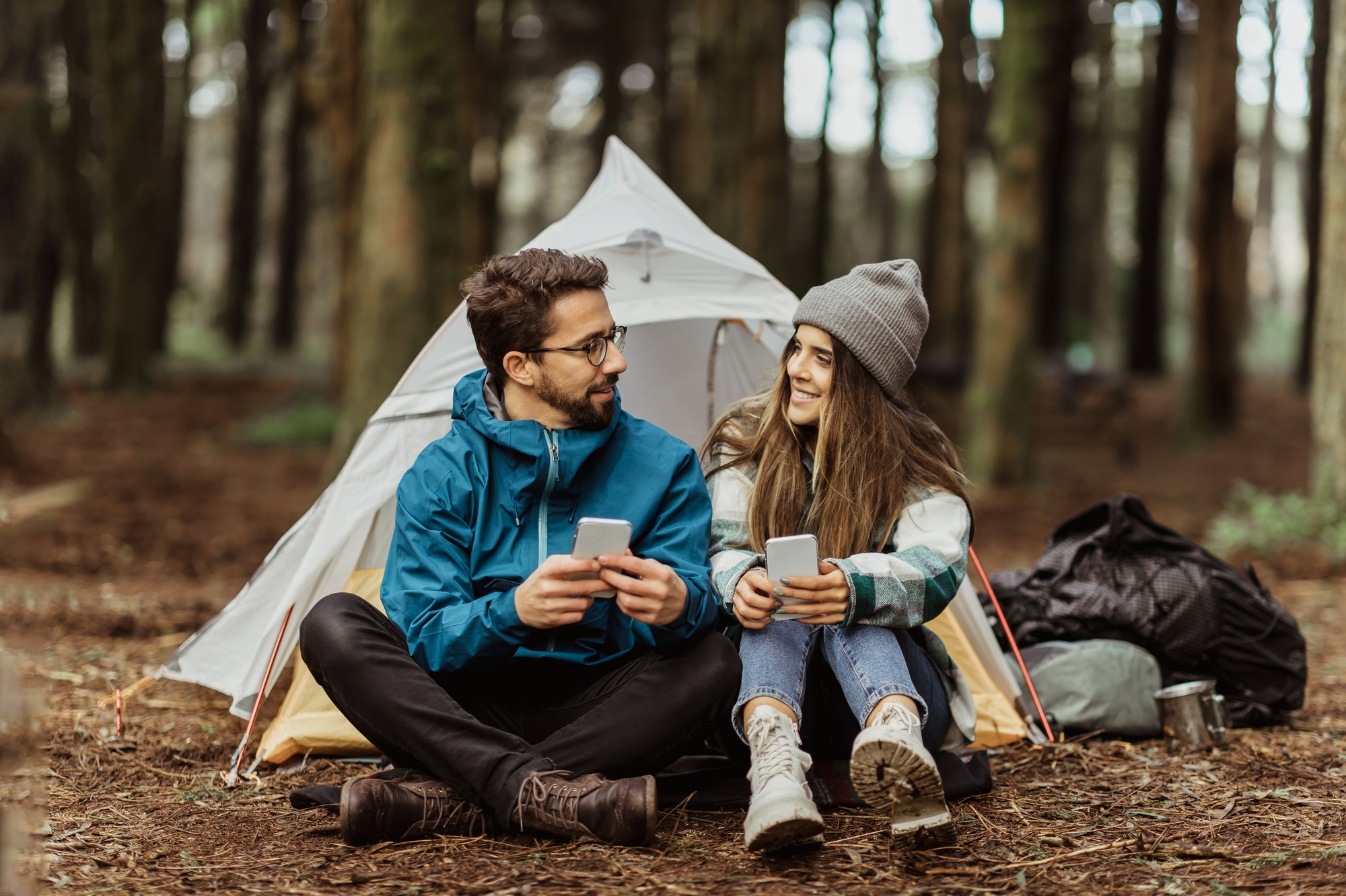 cheerful-young-european-man-and-female-tourists-in-jackets-resting-in-forest-near-tent-typing-on.jpg